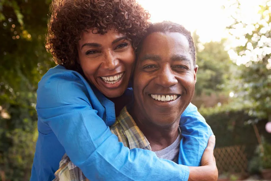 Autoimmune-Doctor A middle-aged black couple outside, the woman embracing the man from behind pressing her cheek to his, both are smiling due to successful Autoimmune Disorder treatment from Delara Tavakoli, M.D. of Tavicare in Chevy Chase.