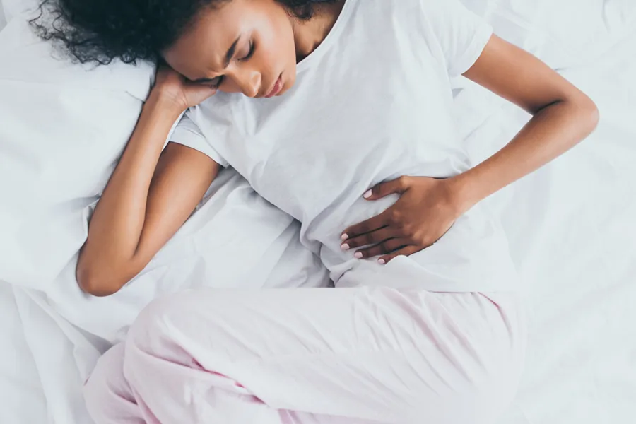 Crohns-Disease-Clinic A dark-skinned woman in white clothes lays on her bed clutching her middle before getting treatment for Crohn's Disease from Delara Tavakoli, M.D. of Tavicare in Chevy Chase.