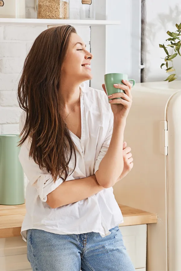 Crohns-Disease-Treatment A woman with a white blouse an jeans drinking tea in her kitchen, smiling after getting treatment for Crohn's Disease from Delara Tavakoli, M.D. of Tavicare in Chevy Chase.