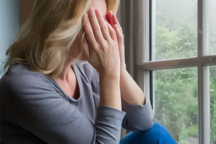 Graves-Disease-Clinic A blonde woman in a gray shirt sits in front of a window on a rainy day covering her eyes with her hands. Get treatment for Grave's Disease from Delara Tavakoli, M.D. of Tavicare in Chevy Chase.