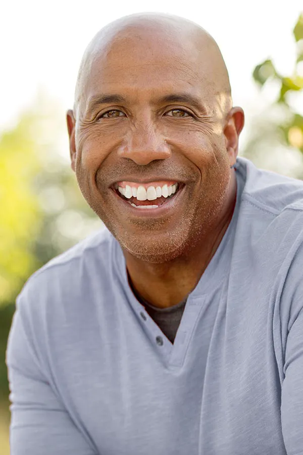 Healthy-Aging-Peptides-Treatment A middle-aged black man in a gray v-neck t-shirt sits outside smiling, happy with the healthy aging peptide treatments from Delara Tavakoli, M.D. of Tavicare in Chevy Chase