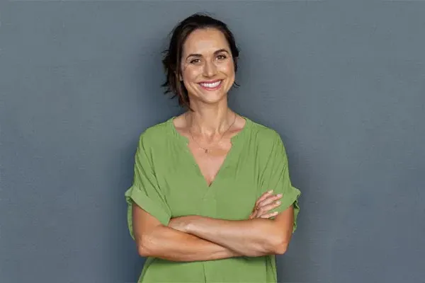 Hormones-for-Women-Doctor A woman in a green shirt stands smiling against a gray-blue wall, pleased with her hormone therapy from Delara Tavakoli, M.D. of Tavicare in Chevy Chase.