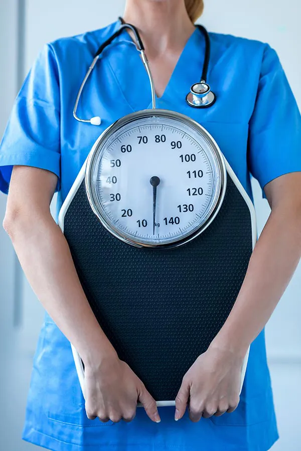 Medical-Weight-Loss-Treatment A nurse in blue scrubs with a stethoscope hanging around her neck holds a scale, representing weight loss treatments from Delara Tavakoli, M.D. of Tavicare in Chevy Chase.