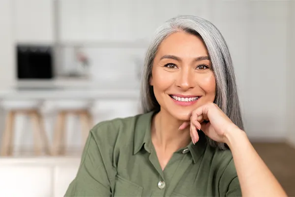 Menopause-Clinic An asian woman with long gray hair and a green shirt sitting and smiling on her couch, happy with her menopause care from [NAME} in Chevy Chase.