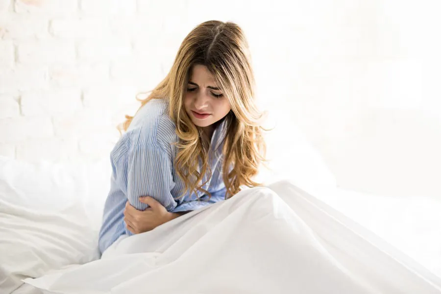 PMS-IV-Relief-Clinic A woman with long blonde hair sits in her bed holding her stomach in pain, considering getting IV care for PMS from Delara Tavakoli, M.D. of Tavicare in Chevy Chase.