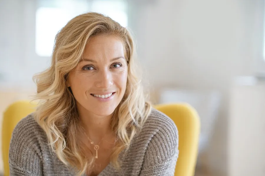 Perimenopause-Doctor A blonde woman wearing a a gray sweater sits inside in a yellow chair, smiling and happy with her perimenopause treatment from Delara Tavakoli, M.D. of Tavicare in Chevy Chase.