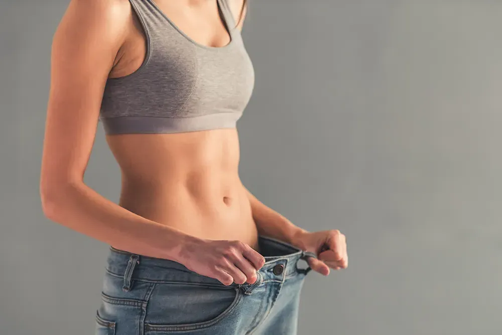 Tesamorelin-Peptide-Clinic A woman pulls too-big jeans away from her waist to show weight loss. Schedule Tesamorelin peptide treatment from Delara Tavakoli, M.D. of Tavicare in Chevy Chase.